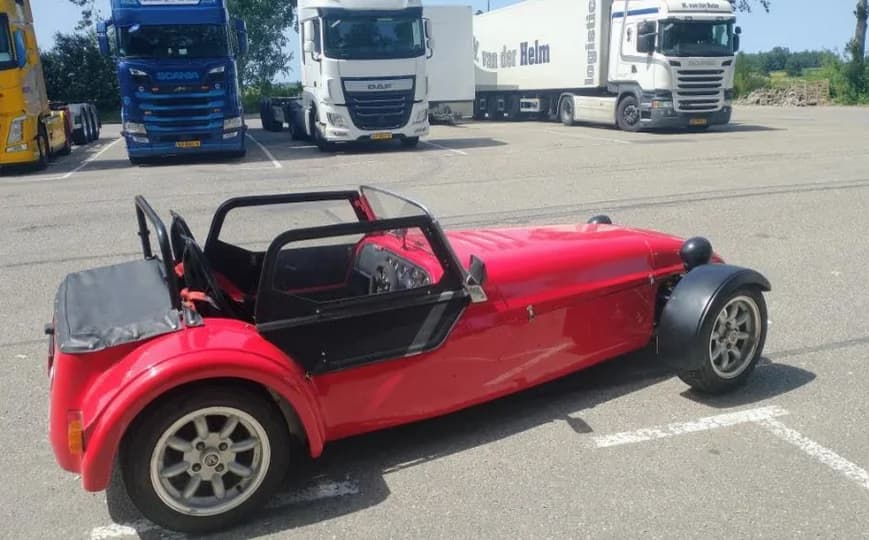 De Westfield zoals hij reed vóór de restauratie: rode widebody op een parkeerplaats tussen vrachtwagens.