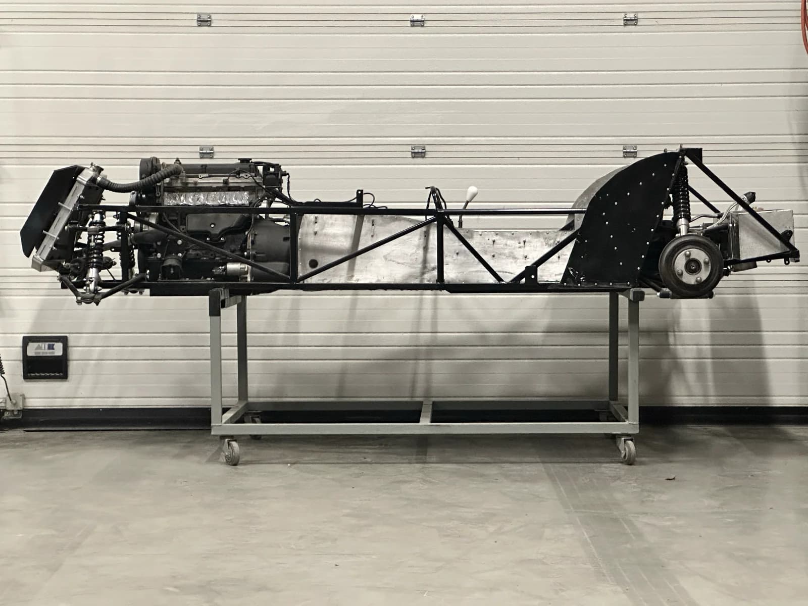 Naakte Westfield chassis op een rolstand, zijaanzicht vanaf links. Motor gemonteerd, buizenwerk en ophanging duidelijk zichtbaar tegen een lichte werkplaatswand.