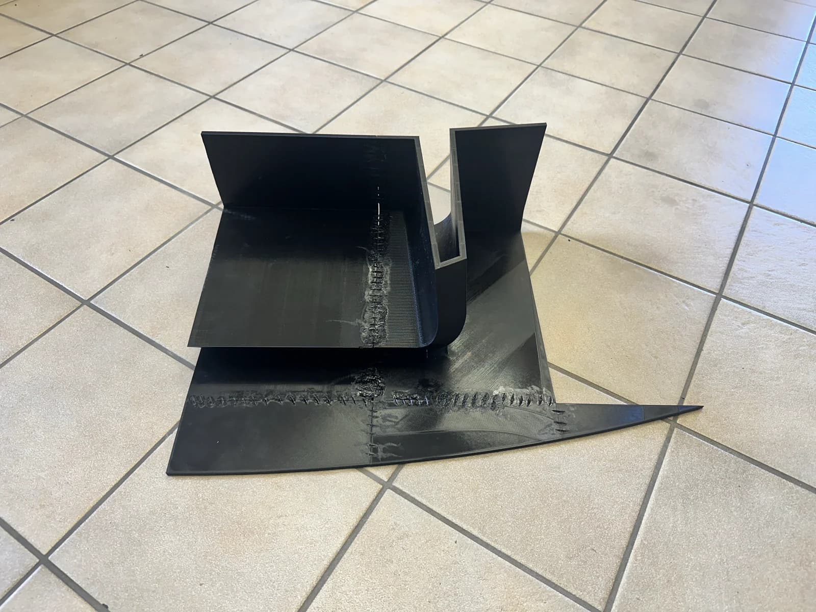 Twee zwarte geprinte body-stukken aan elkaar gesmolten op een lichte tegelvloer, met de plastic welder. De smeltnaden zijn duidelijk zichtbaar als donkere sporen op het gladde oppervlak.