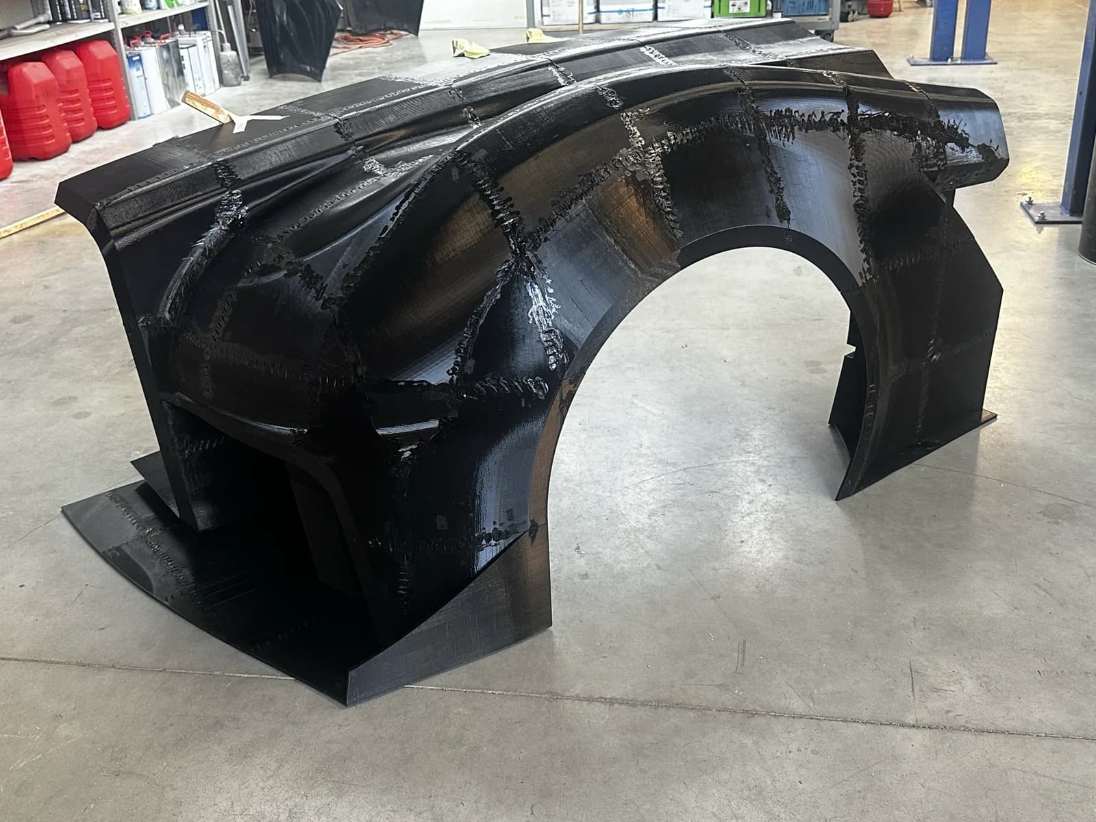 Grote samenhangende body-sectie op een garagevloer: rechter-achterkwart met wielkast en flank. Autolift en werkplaatsomgeving op de achtergrond geven de schaal aan.
